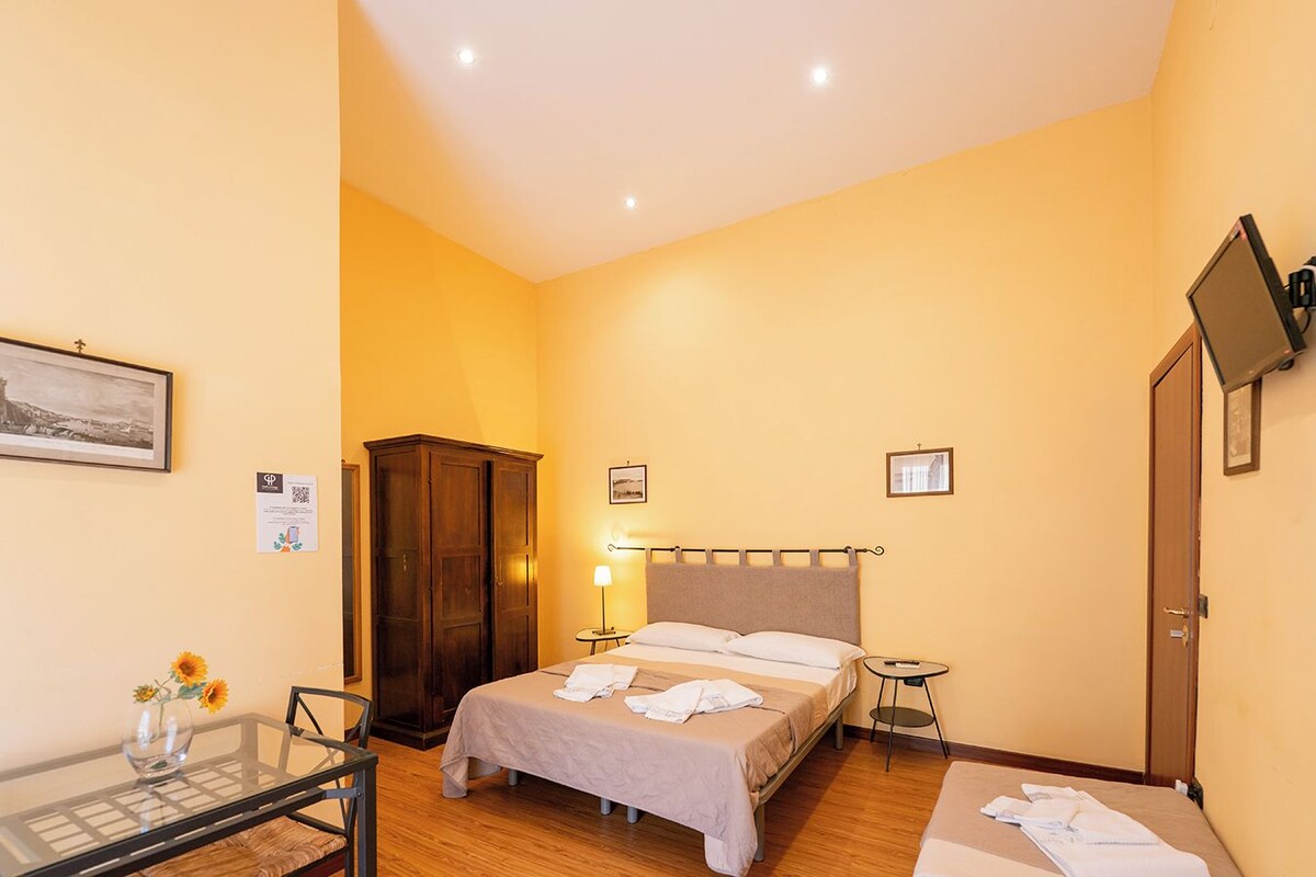 A bedroom with a bed, a wardrobe, a desk with a chair, a lamp, and a TV on the wall.