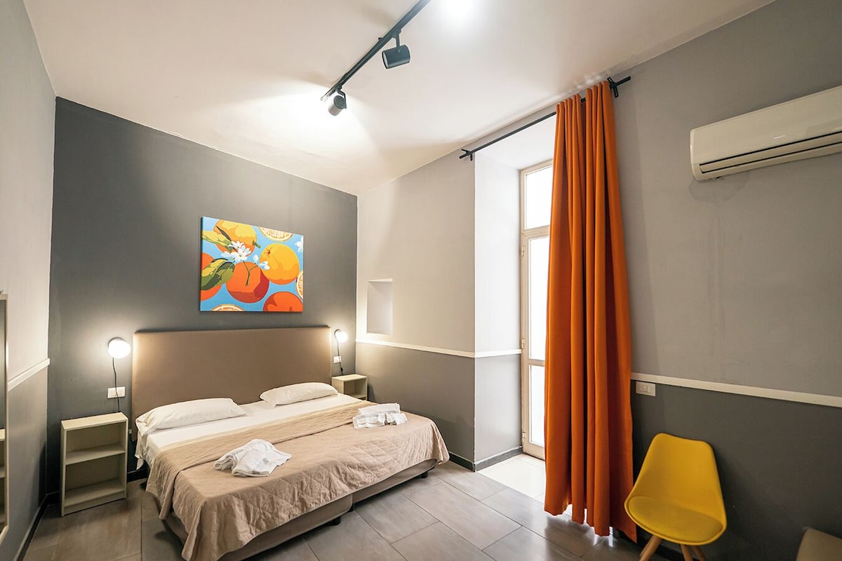 A hotel room with a bed, bedside lamps, a painting, a window with curtains, and an air conditioning unit.