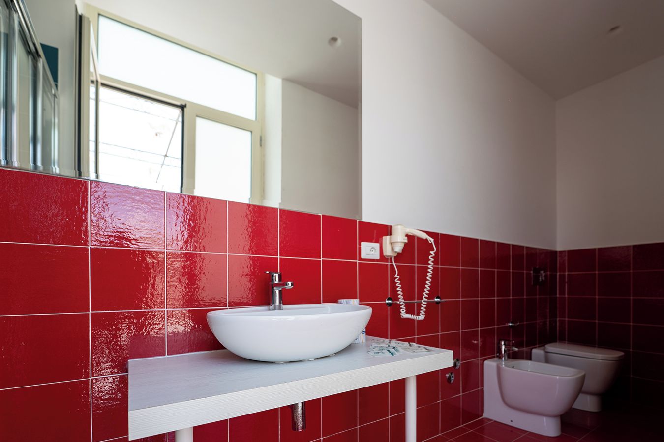 Une salle de bain avec des murs recouverts de carreaux rouges, un lavabo blanc, un miroir, une douchette à main et des toilettes.