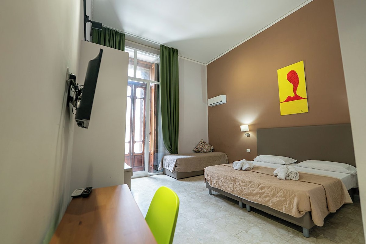 A hotel room with two beds, a desk, a chair, a window with curtains, and a wall-mounted air conditioner.