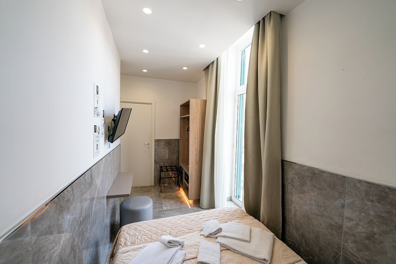 A modern hotel room with a bed, a small seating area, a TV mounted on the wall, and a window with curtains.