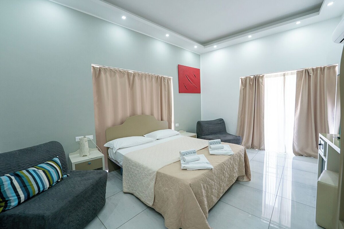 A modern hotel room with a large bed, a desk, a chair, and a sofa.