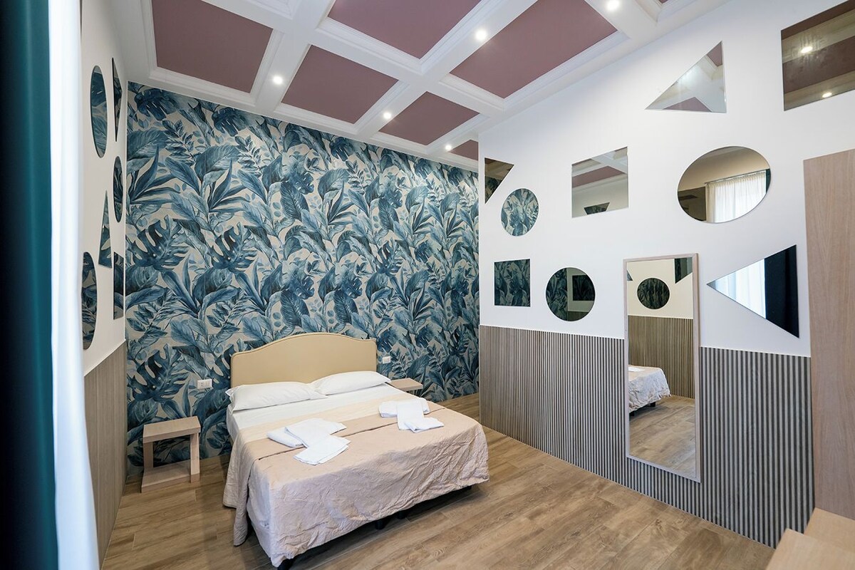 A bedroom with a bed, a nightstand, a mirror, and a wall with circular and rectangular mirrors.