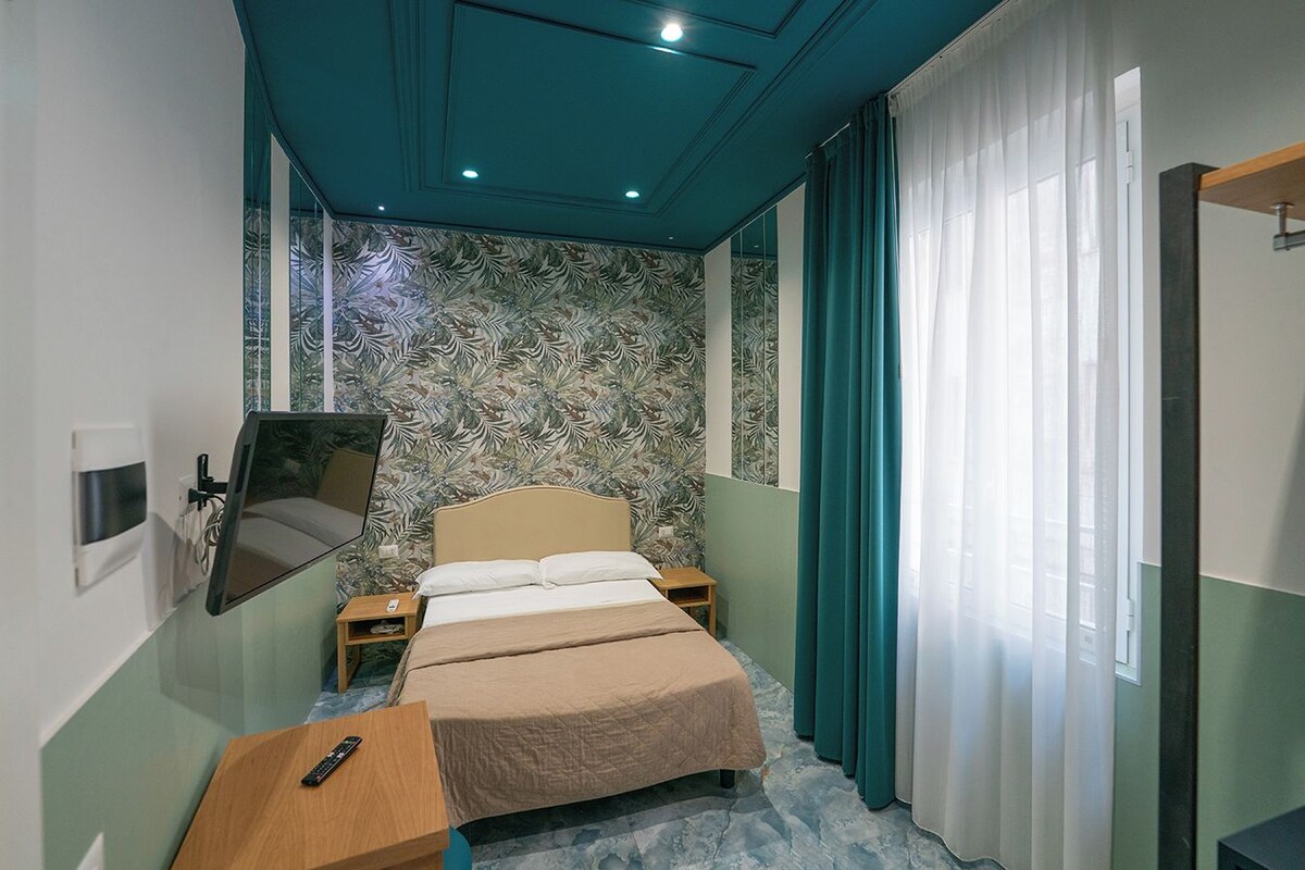 Hotelkamer met een bed, nachtkastje, spiegel, televisie en raam met gordijnen.