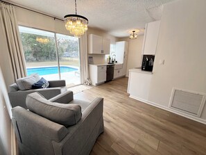 Ein modernes Wohnzimmer mit grauem Sofa, einer Chandelier und einer Küche mit weißen Schränken, mit Blick auf einen Swimmingpool.