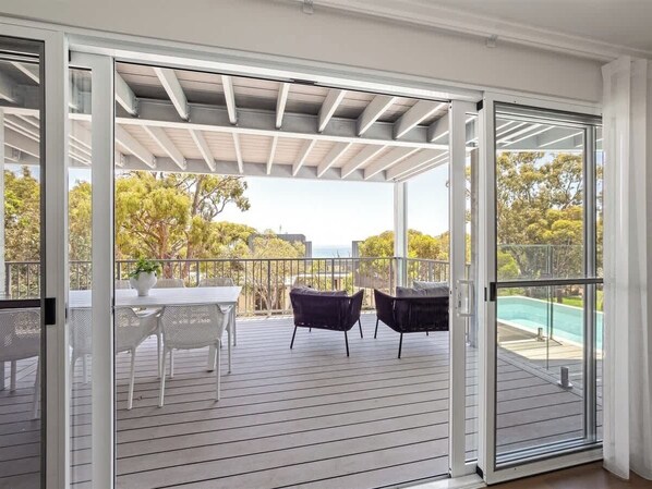 A covered outdoor area with a table and chairs, a pool in the background, and a view of trees.
