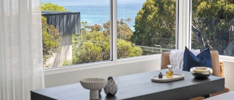Habitació moderna amb un gran finestral que ofereix vistes a la costa, una taula de menjador amb un bol i una ampolla, i un llit amb un coixí.