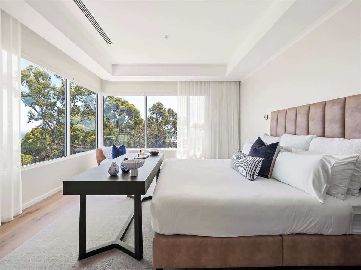Une chambre moderne avec un grand lit, une table de chevet et une vue sur les arbres à travers de grandes fenêtres.