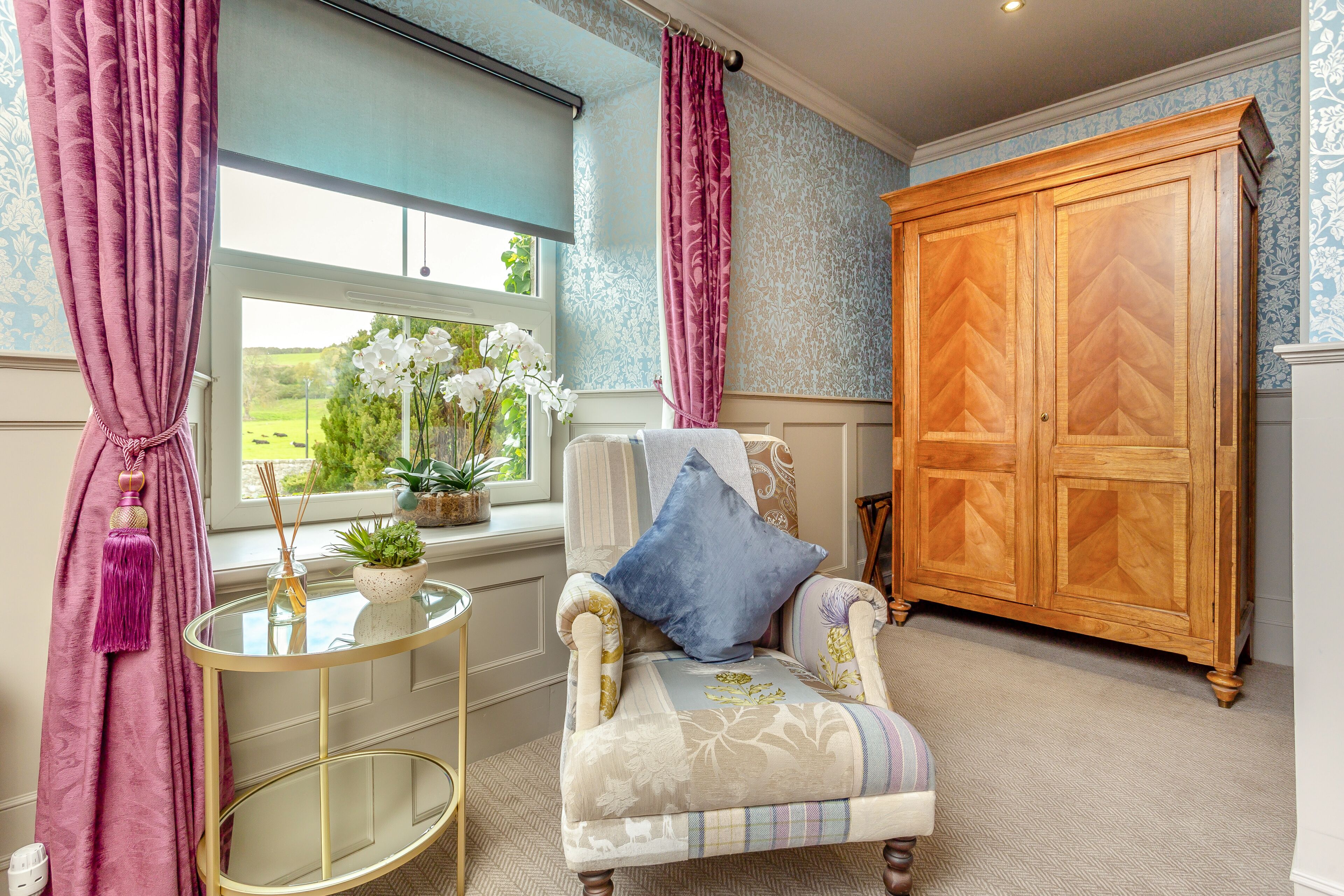A room with a large wooden wardrobe, a round glass table with a plant, and a chair with a blue pillow.
