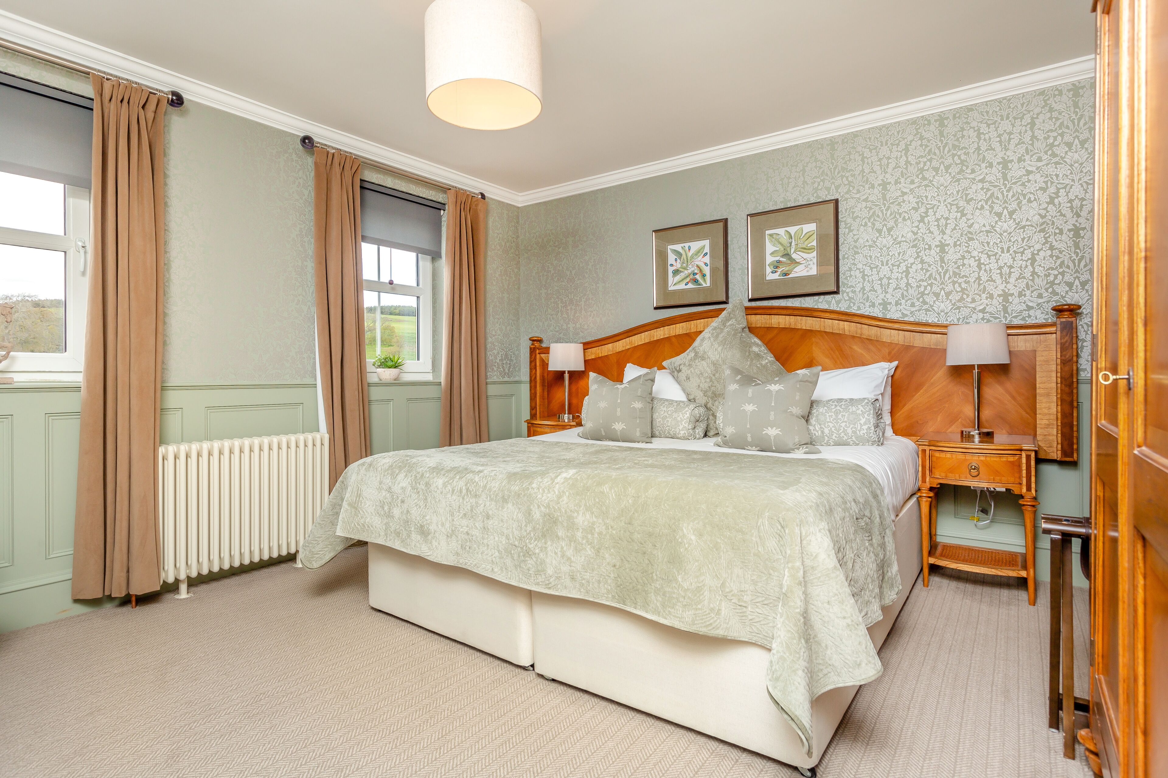 Une chambre à coucher avec un grand lit, deux tables de chevet, une commode, un radiateur et une fenêtre avec des rideaux.