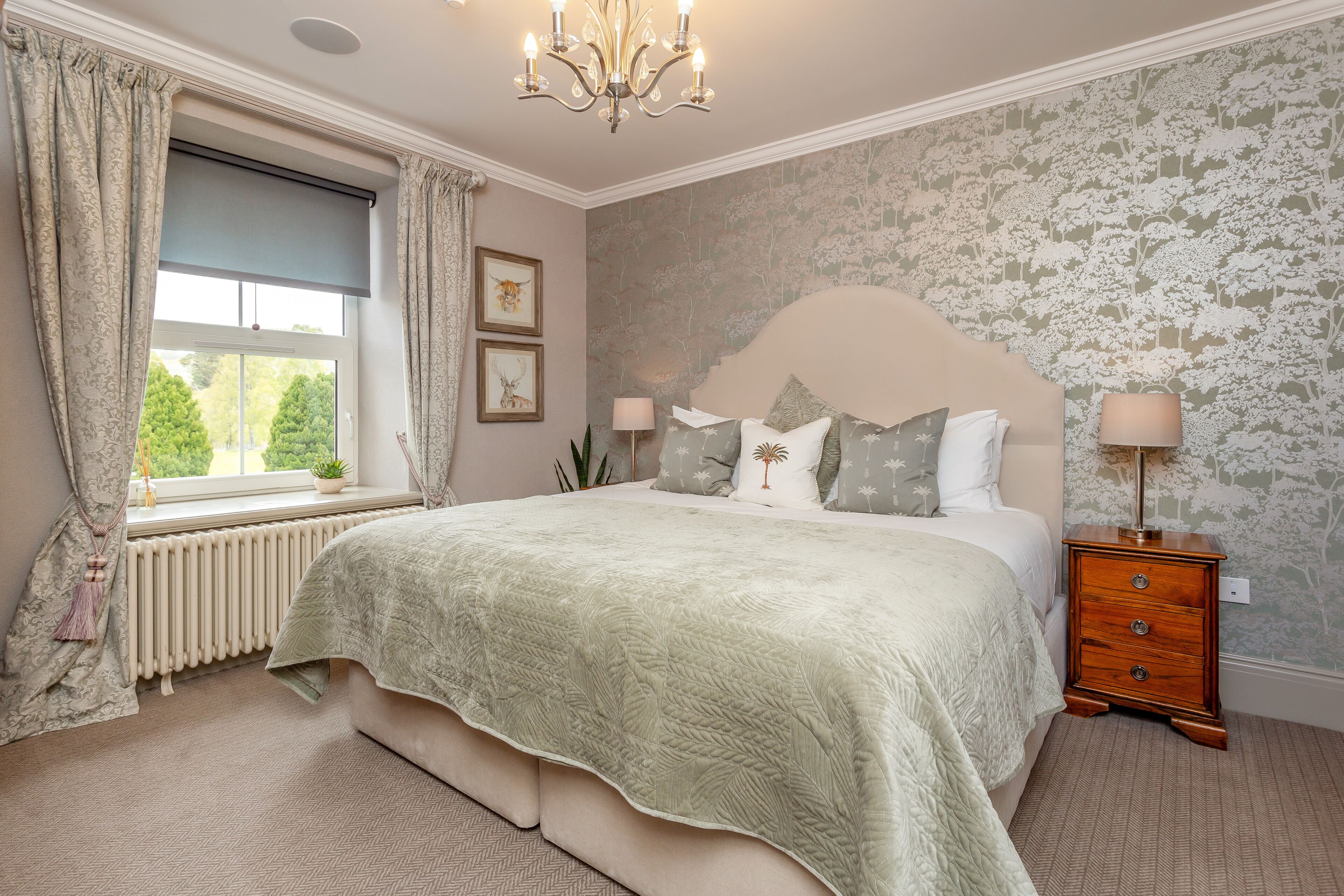 A bedroom with a large bed, a nightstand, a radiator, a window with curtains, and a wall with floral wallpaper.