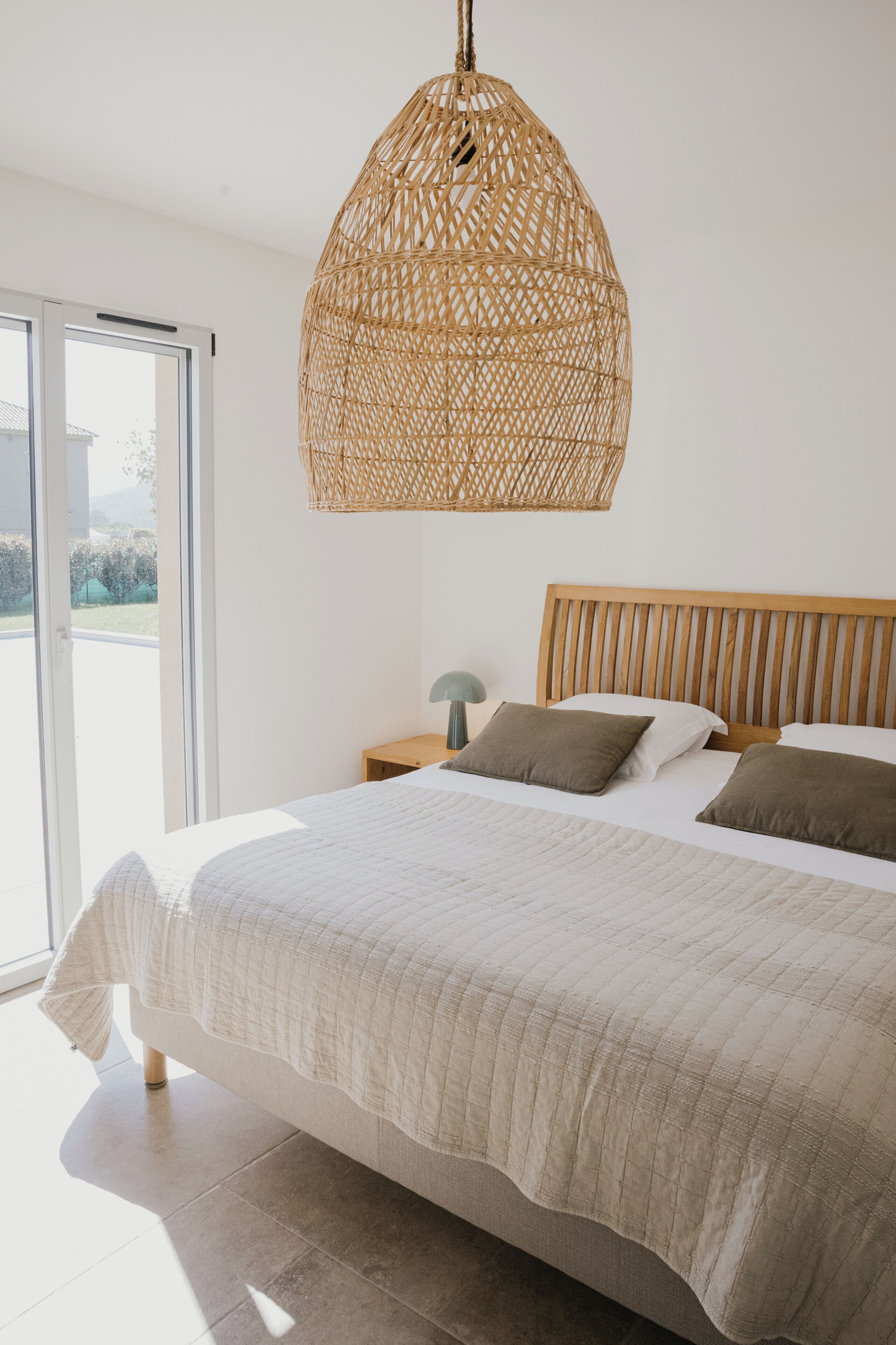 Quarto com uma cama grande, cabeceira de madeira, luminária pendente de vime e vista através de uma porta de vidro.