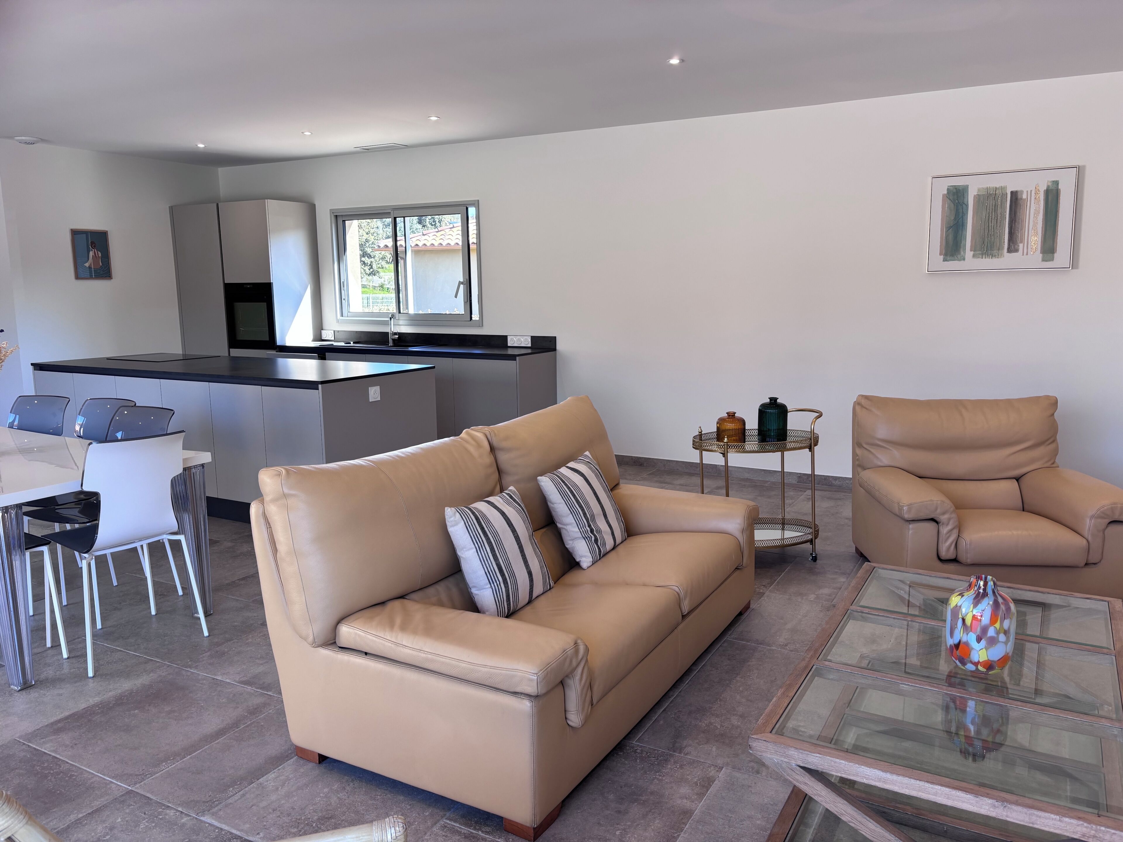 A modern living room with a beige sofa, a glass coffee table, and a dining area with chairs and a kitchen in the background.