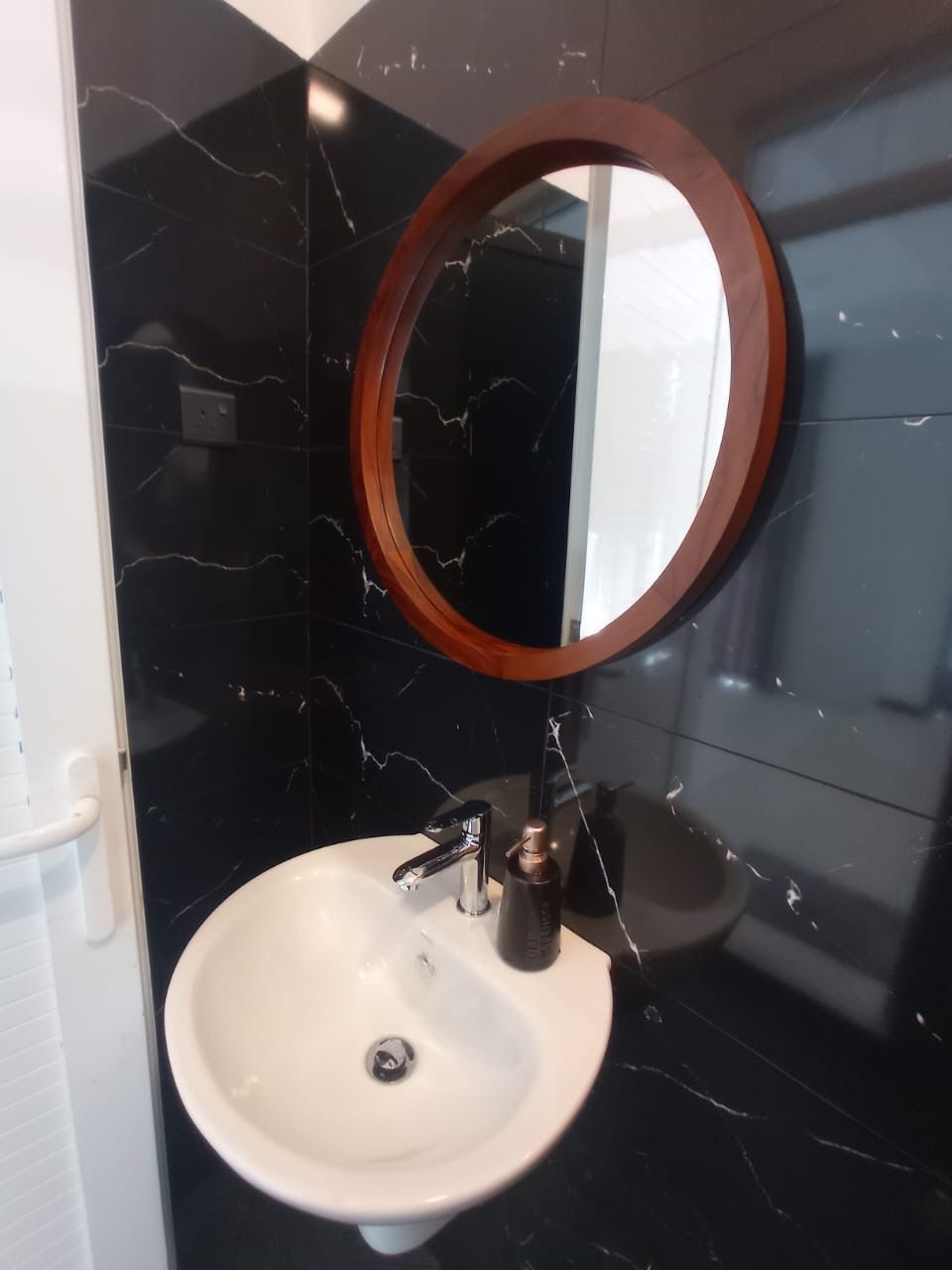 Ein Badezimmer mit weißem Waschbecken, einem runden Spiegel mit Holzrahmen und schwarzen Marmorwänden.