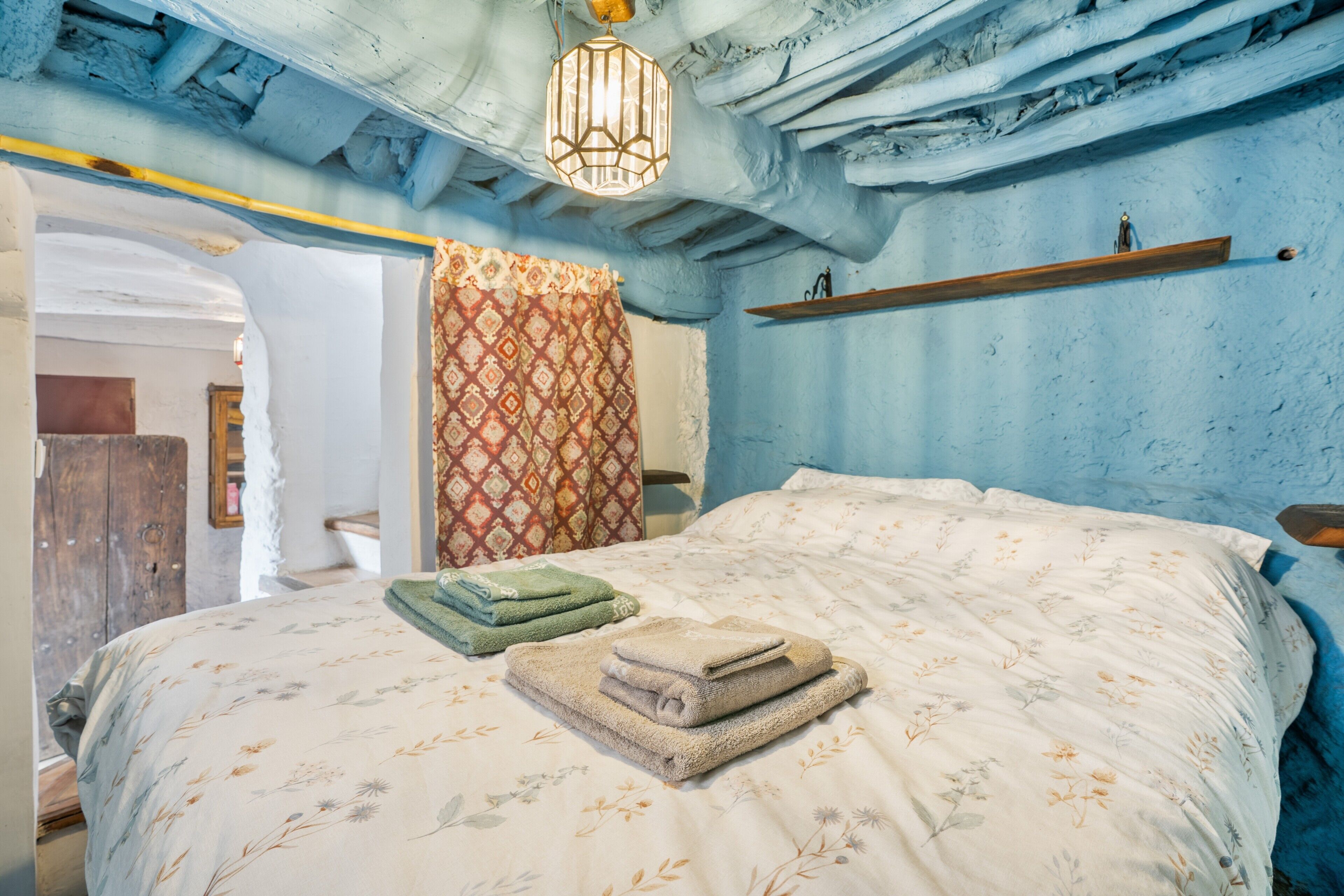 Een slaapkamer met een bed, een behang met patroon, een hanglamp en een houten plank.