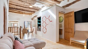 Una sala de estar con un sofá, una mesa redonda y una silla de madera. Hay un televisor montado en la pared y una puerta de madera.