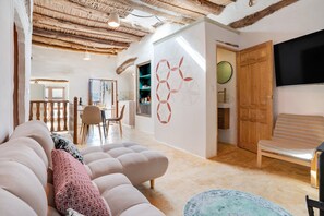Un salon avec un canapé, une table ronde et une chaise en bois. Un téléviseur est fixé au mur et il y a une porte en bois.