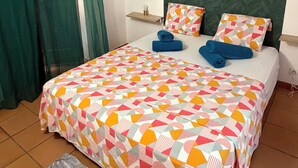 Een netjes opgemaakt bed met een kleurrijk, geometrisch beddegoed, een klein nachtkastje met een lamp, en een groen gordijn.