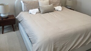 En snyggt bäddad säng med ett beige täcke och kuddar, en sänglampa och ett litet träbord vid sängen med en lampa på.