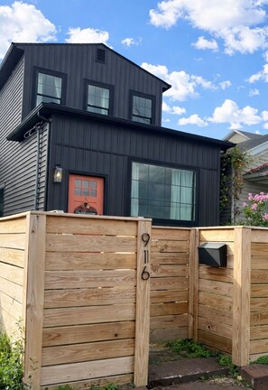 Une maison moderne à deux étages, dotée d’une façade sombre, de grandes fenêtres et d’une clôture en bois avec une boîte aux lettres.