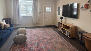 Ett vardagsrum med en stor TV, en bokhylla, en blå soffa och en mönstrad matta.