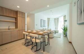 Uma cozinha moderna com armários de madeira, um espaço de jantar com mesa posta para quatro pessoas e uma sala de estar com sofá e planta em vaso.