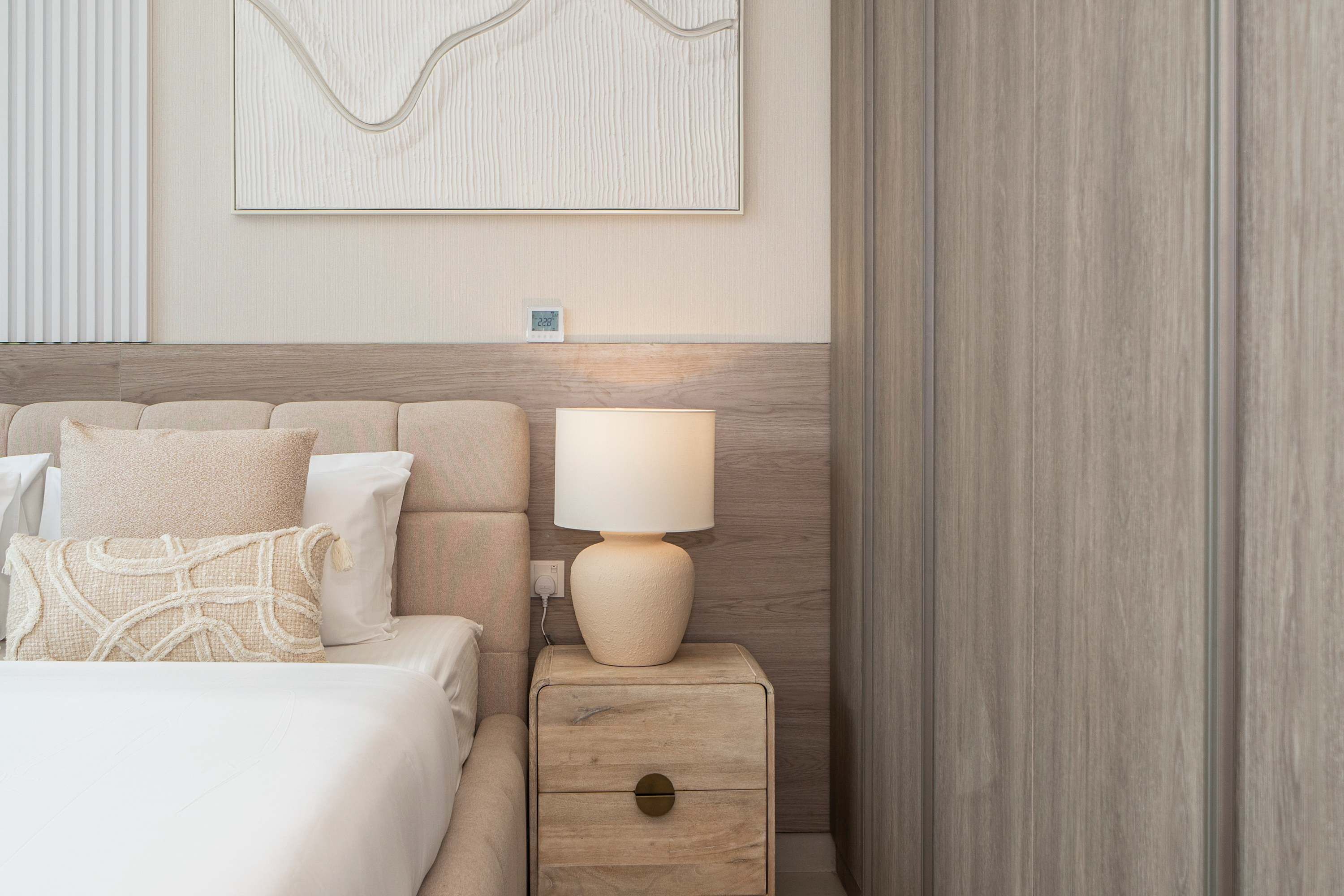 A neatly made bed with a headboard, a bedside lamp, a nightstand with a drawer, and a wall-mounted clock.