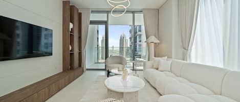 A modern living room with a large flat-screen TV, a white sofa, a round coffee table, and a view of tall buildings through large windows.