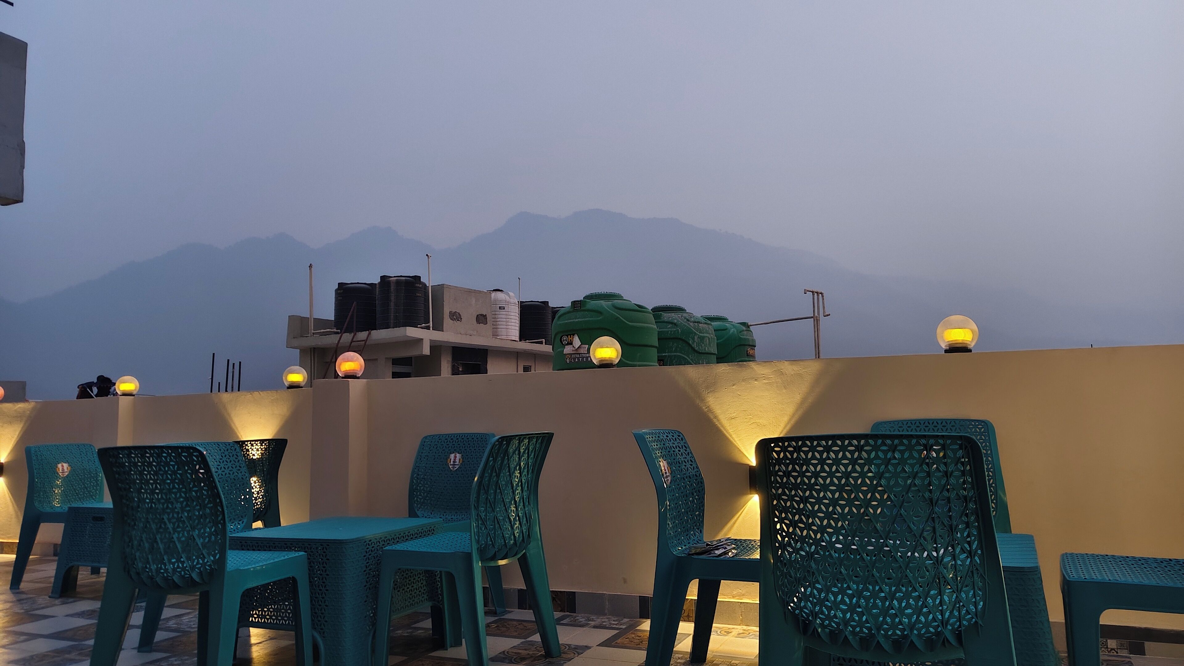 A rooftop terrace with teal chairs and tables, water tanks, and a view of distant mountains.