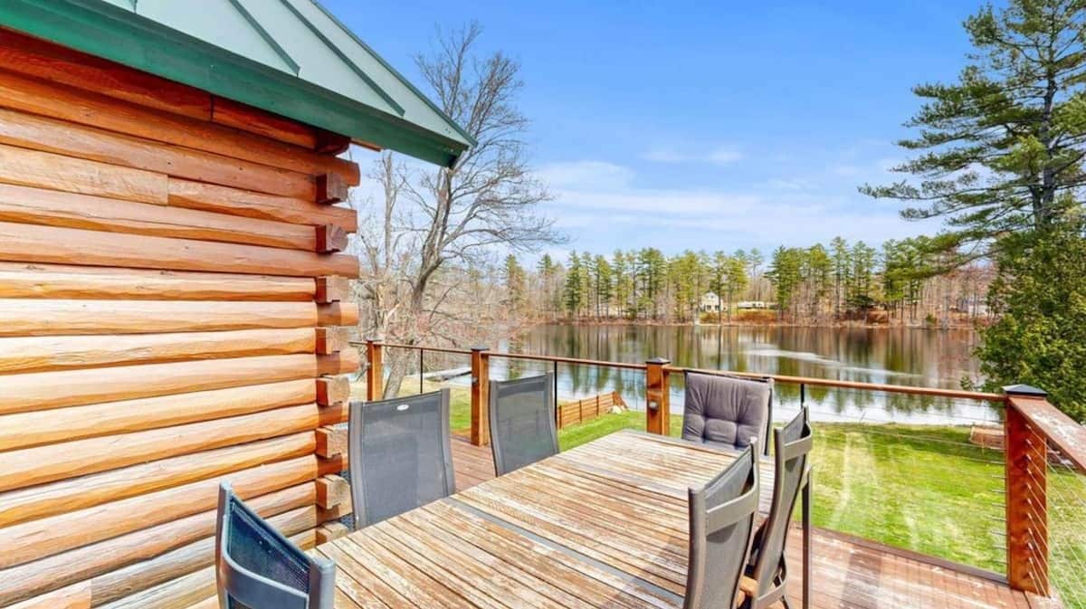 A wooden deck with a table and chairs, overlooking a lake and forest.