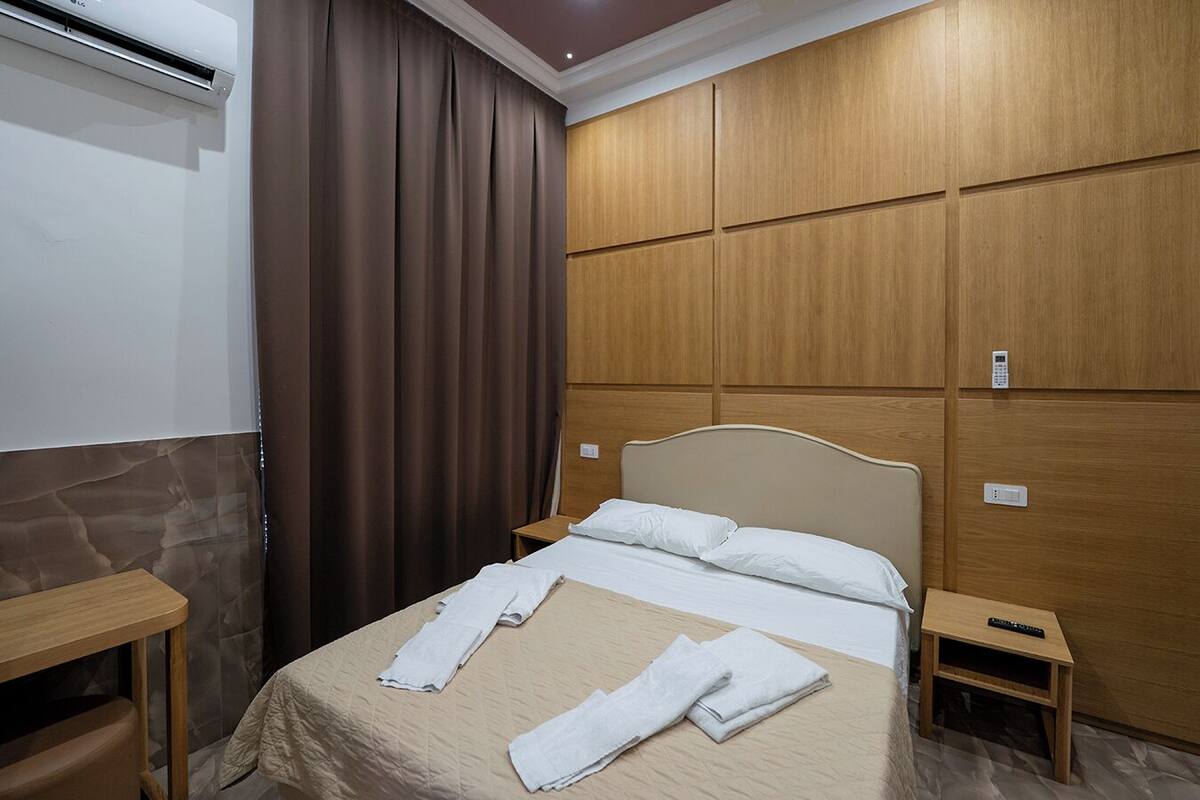 A hotel room with a bed, wooden panel walls, a curtain, a remote control, and a wall-mounted air conditioner.