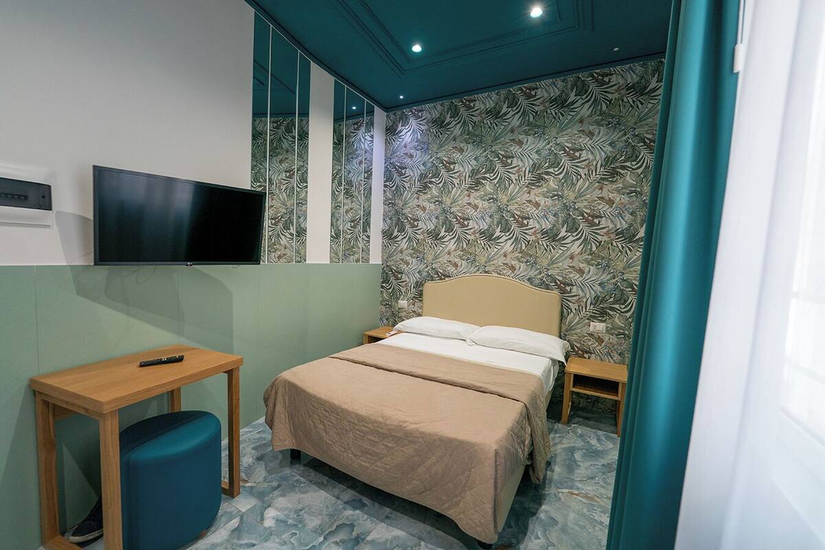A bedroom with a bed, a TV mounted on the wall, a small table, and a trash bin.