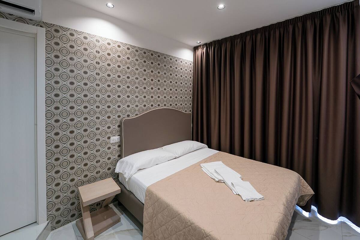 A modern hotel room with a patterned wallpaper, a large bed with a headboard, a bedside table, and a window with curtains.