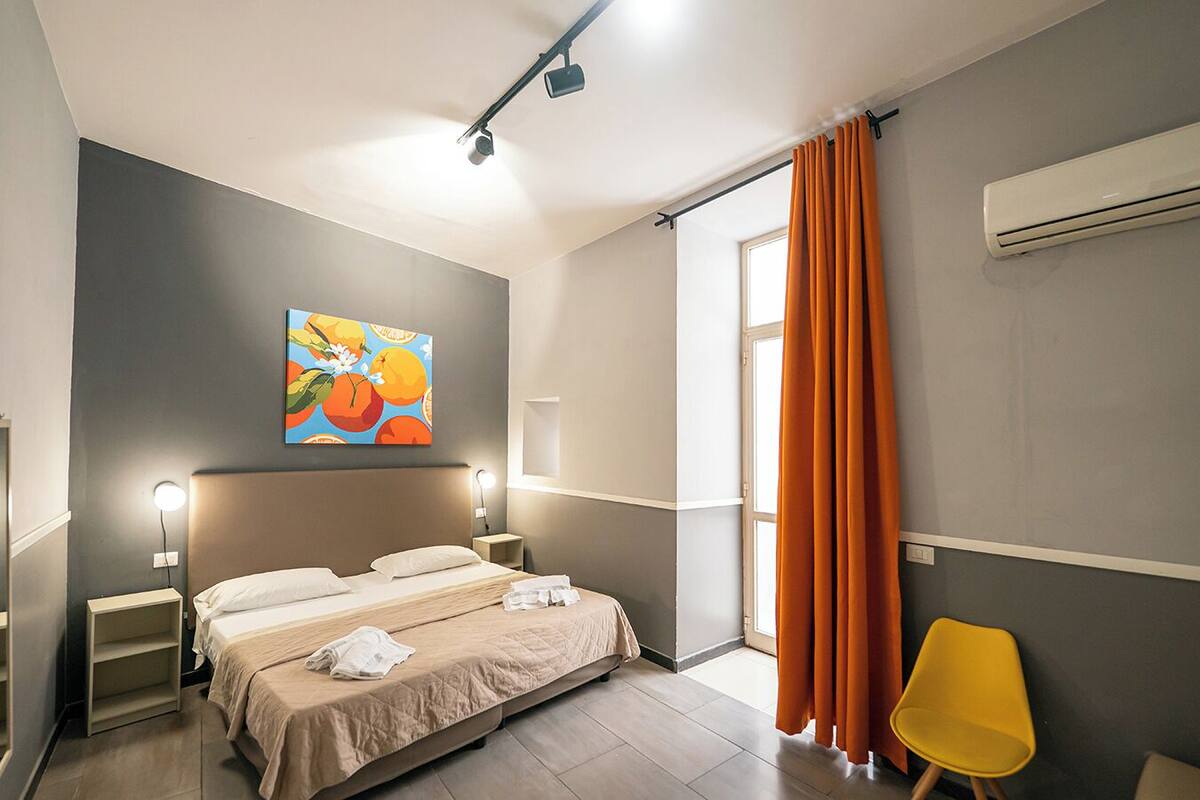 A hotel room with a bed, bedside lamps, a painting, a window with curtains, and an air conditioning unit.