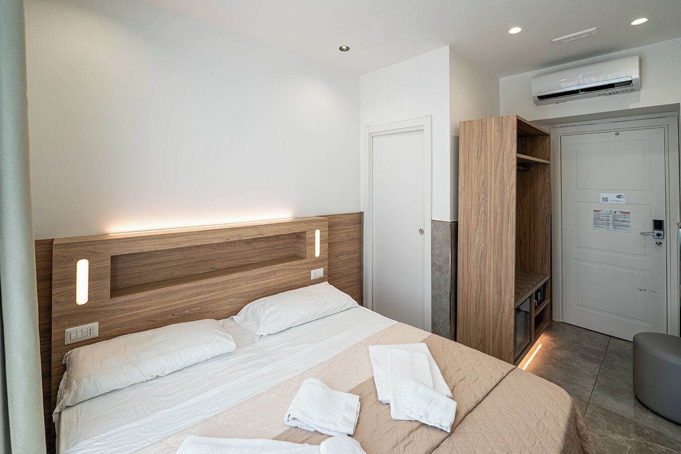 A modern hotel room with a wooden headboard, a bed with white linens, a bedside table, an air conditioning unit, and a wardrobe.