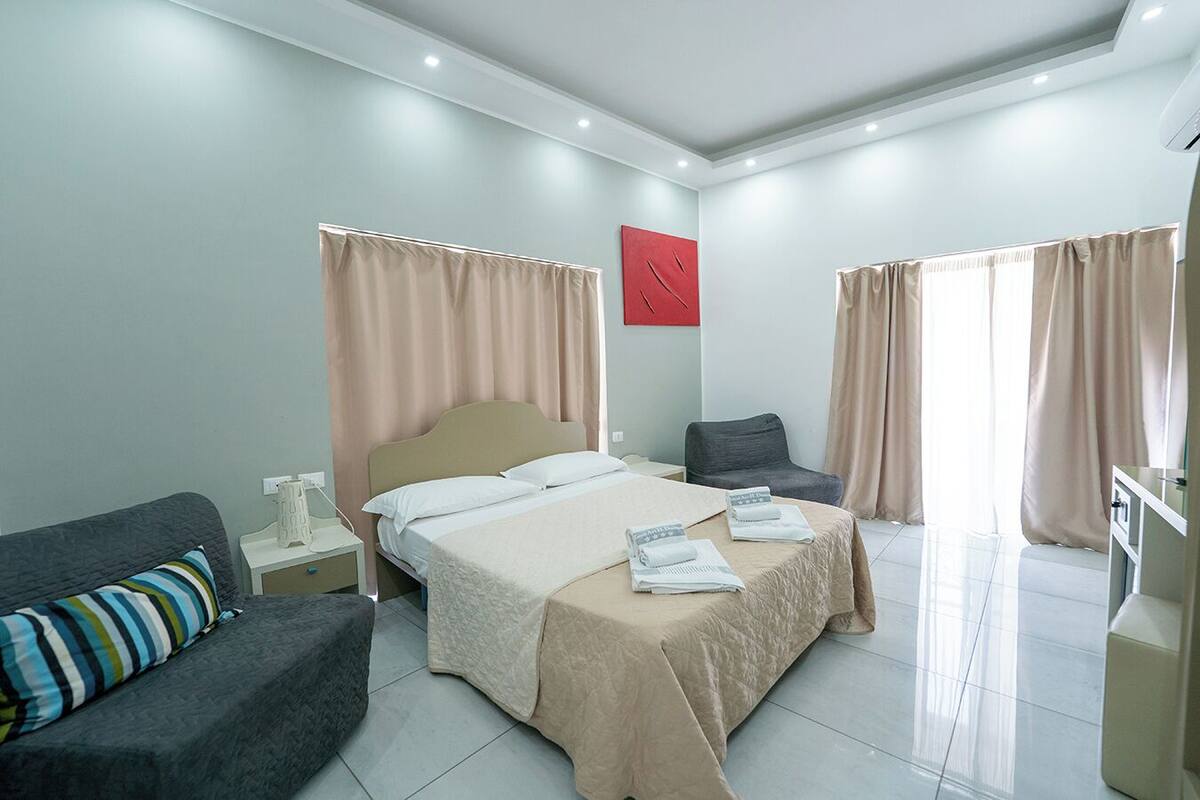 A modern hotel room with a large bed, a desk, a chair, and a sofa.