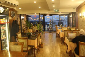 A restaurant with wooden furniture, a Christmas tree, and a view of the city through the window. - GROSS HOTEL (Istanbul)