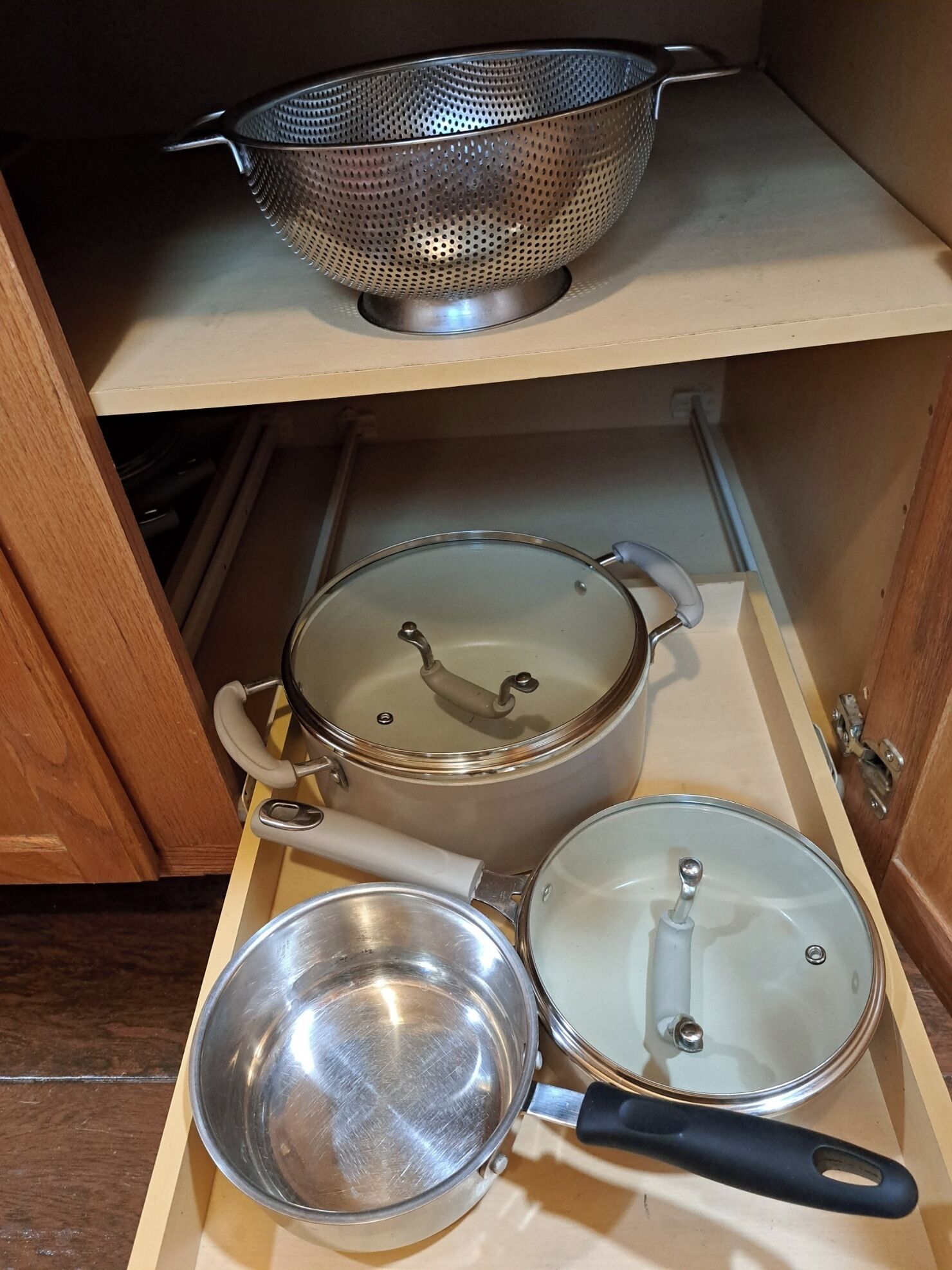 Un armario de cocina con un colador de metal, una olla de acero inoxidable con tapa y una sartén con tapa.