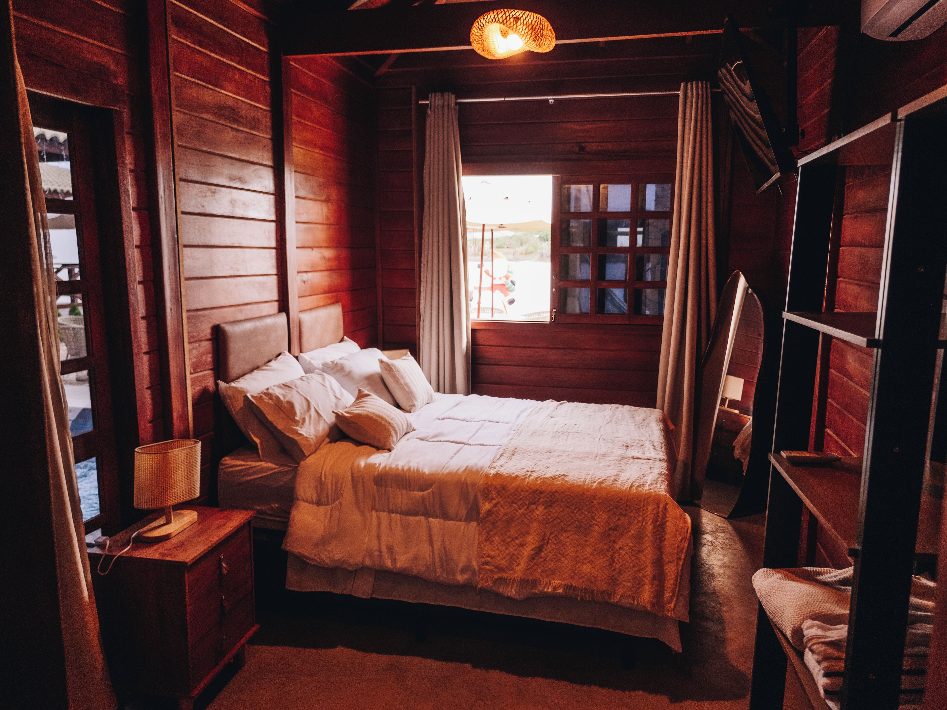 A wooden cabin bedroom with a bed, nightstand, lamp, and a view of the outside through a window.
