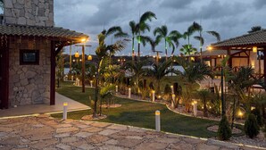 A stone building with a tiled roof, surrounded by palm trees and a well-lit garden at dusk. - Lagoon Paradise (Fortim)