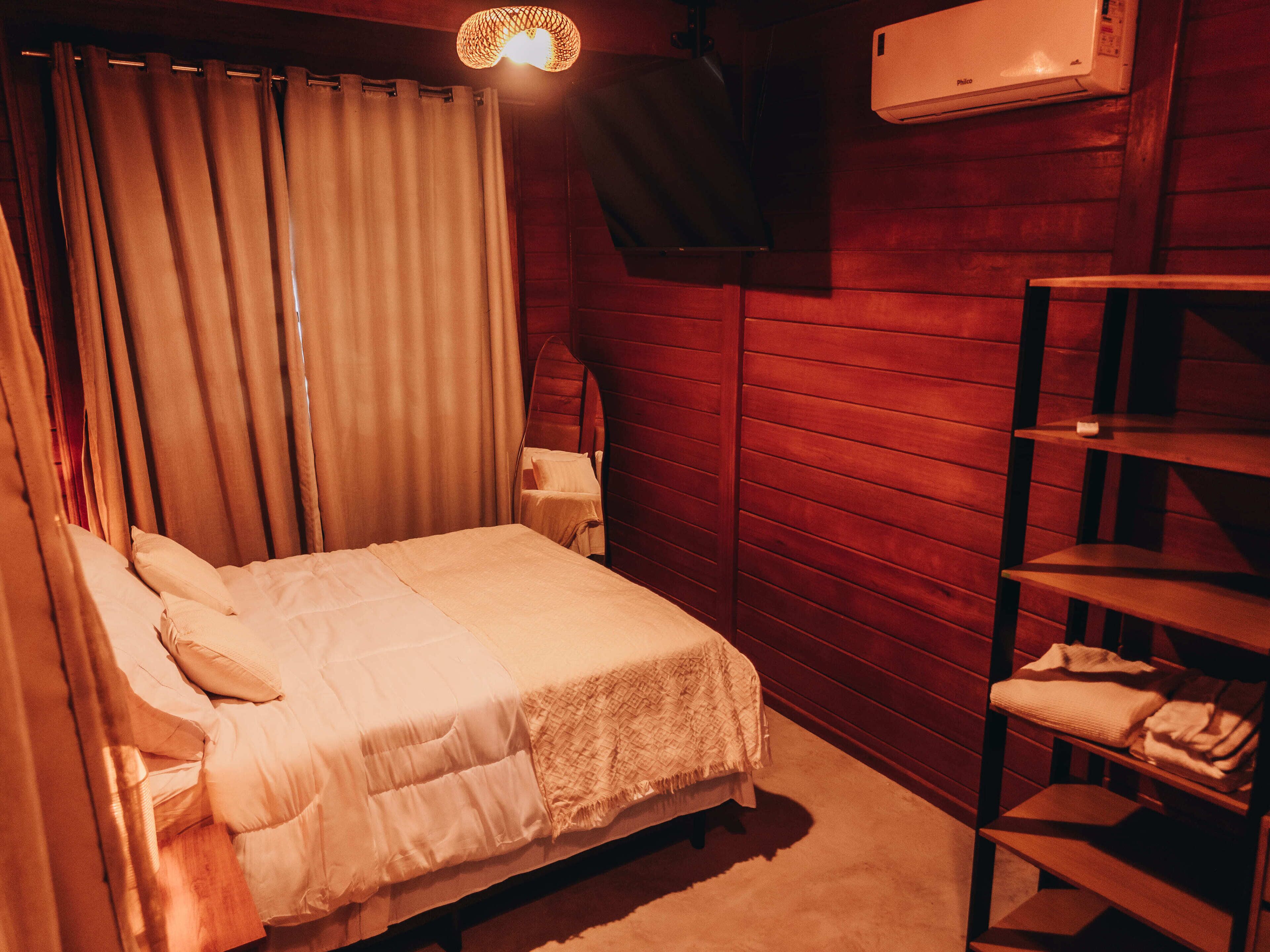Una habitación con cama, repisas con toallas dobladas, aire acondicionado de pared y cortina.