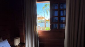 Quarto de madeira com cama, cortinas e janela com vista para a praia.