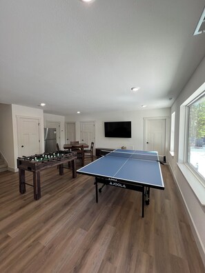 A room with a ping pong table, foosball table, and a flat-screen TV mounted on the wall. - 9BR Theater Room Retreat: Dock Slip in cove, Sleeps 30! EV charger! (Lafollette)