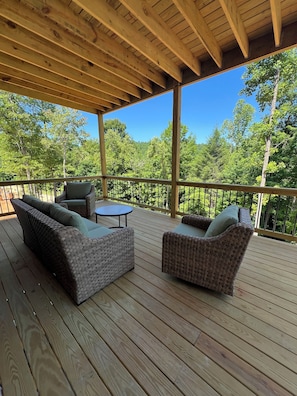 Une terrasse en bois, agrĂ©mentĂ©e de meubles en osier et dâune table, entourĂ©e dâune rambarde en mĂ©tal, et offrant une vue sur une zone boisĂ©e.