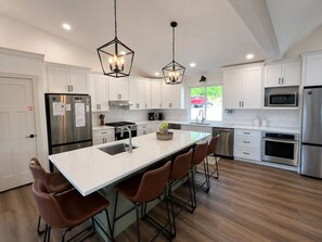 Una cocina moderna con armarios blancos, electrodomésticos de acero inoxidable, una isla central con taburetes e iluminación colgante.