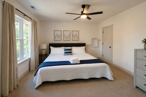 Quarto com cama, ventoinha no teto, mesa de cabeceira, luminária, cômoda e janela com cortinas.