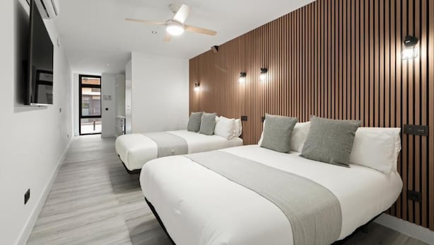 Habitación de hotel moderna con dos camas, un revestimiento de pared de madera, ventilador de techo y vista al pasillo.