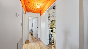 Een gang met houten vloer, een witte deur en een keuken met een bakstenen muur en een hanglamp.