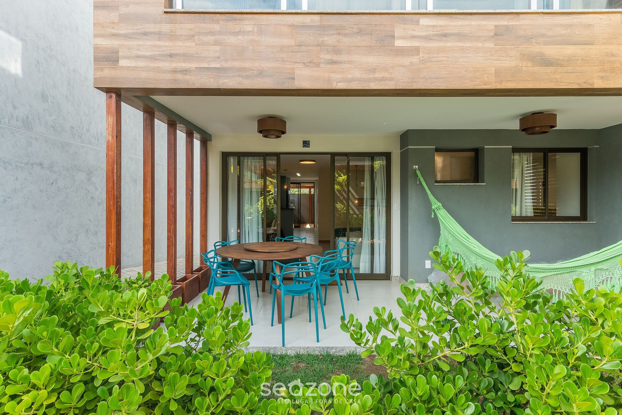 Uma casa moderna com um deck de madeira, uma área de refeições com cadeiras azuis e uma rede.