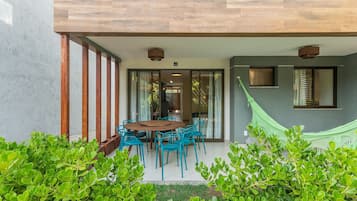 Uma casa moderna com um deck de madeira, uma área de refeições com cadeiras azuis e uma rede.